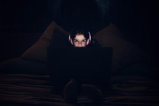 little girl sitting on the bed between cushions while playing or watching a movie on a computer in the darkness, has a pink headset, child and technology concept, copy space for text
