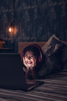 vertical photo of a happy little girl lying in bed smiling while watching a movie on a computer in the darkness, has a pink headset, child and technology concept, copy space for text