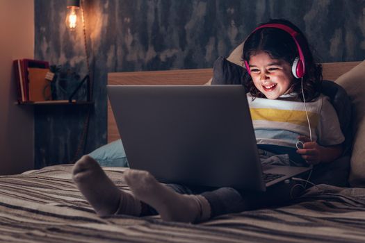 little girl sitting on the bed between cushions laughing while playing or watching a movie on a computer in the darkness, has a pink headset, child and technology concept