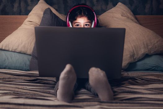 little girl sitting on the bed between cushions focused while playing or watching a movie on a laptop in the darkness, has a pink headset, child and technology concept