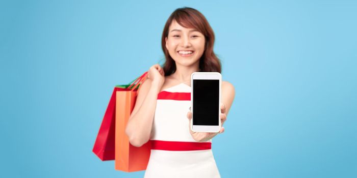 Happy lady with colored paper bags shows smartphone with blank screen and takes order isolated on blue background