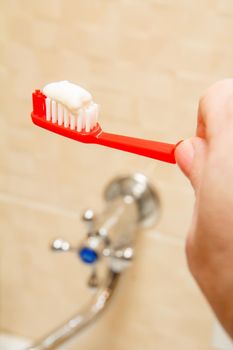 Red toothbrush with white toothpaste in female hands with a water tap on the background.