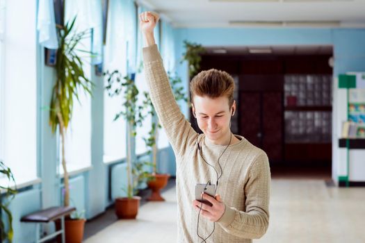 Cheerful male college learner with mobile phone and earphones put up a hand and express victory and happiness