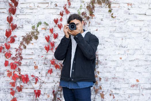 Portrait of young beginner photographer standing near white wall background and shooting someone