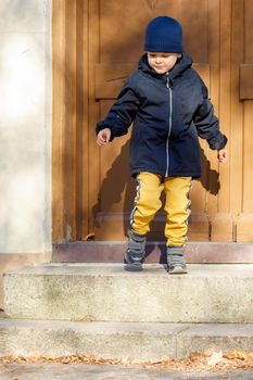 The little boy opens the big brown entrance door. The child leaves the house for an autumn walk.