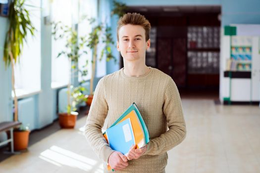 Portrait of attractive cheerful male college student smiling at camera holding books and papers in his hands