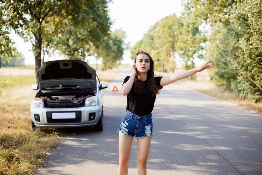 Attractive young student woman tries to stop passerby car and ask for a help because her car has broken