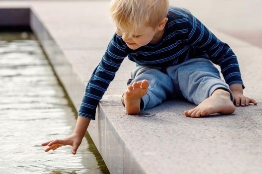 A little boy in the city plays near the fountain with water at summer. Happy boy touching water in fountain.