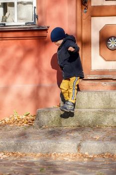 A cute boy in a autumn jacket with a blue hat goes for a walk around the old town. The child is self-climbing the stairs.