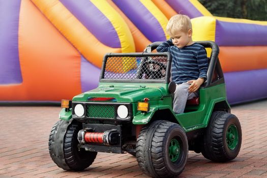 Little child driving green toy car, colored inflatable trampoline in background.