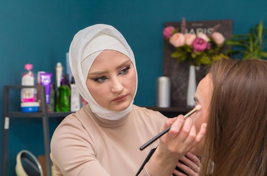 Make-up artist with a brush paints the eyelid of a girl in the salon. Female master makes makeup to a young woman. Business concept - beauty salon, facial skin care, cosmetology.