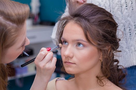 Professional make-up artist with a brush paints the eye of a girl in the salon. Female master makes makeup to a young woman. Business concept - beauty salon, facial skin care, cosmetology.