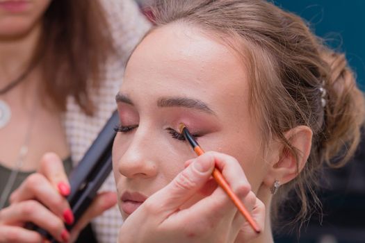 A hand with a brush paints the eyelid of a girl with a large palm. Female master makes makeup to a young woman. Business concept - beauty salon, facial skin care, cosmetology.