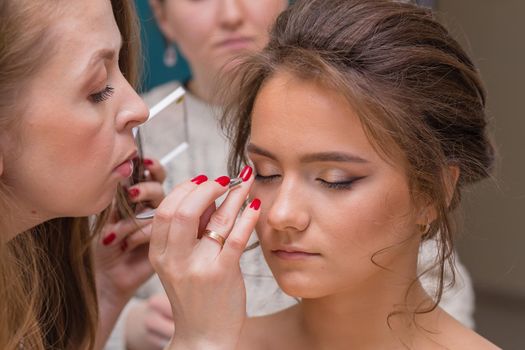 A professional make-up artist corrects shadows on a girl's eyelid with a finger. Female master makes makeup to a young woman. Business concept - beauty salon, facial skin care, cosmetology.