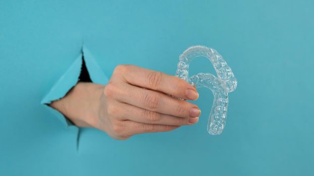 A female hand sticking out of a hole from a blue background holds removable night retainers