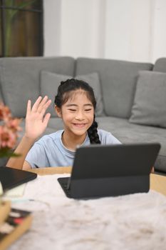 Cute girl having learning online at virtual class on computer tablet while sitting in living room. Concept of Virtual education, homeschooling.
