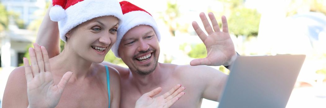 Man and woman in santa hats waving hand at laptop screen on vacation at resort. New year holidays remote congratulation concept