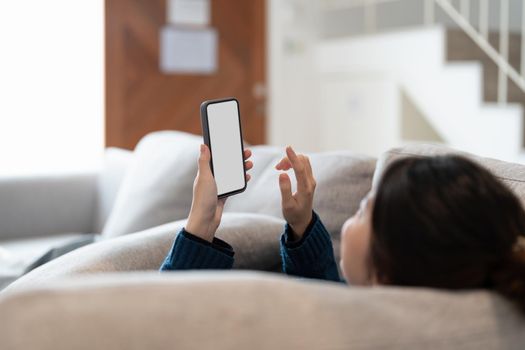 Happy young asian woman lying on sofa at living room and using cell phone at home.
