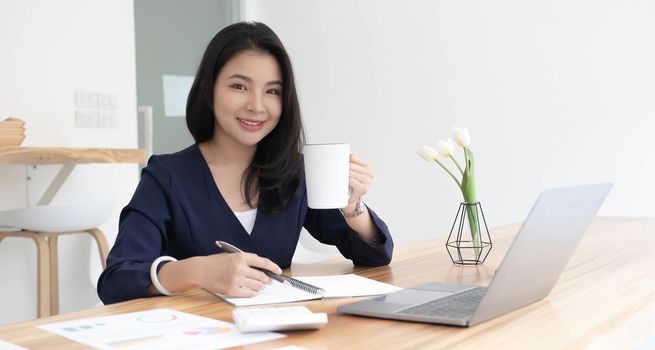 Smiling young Asian businesswoman holding a coffee mug and laptop at the office. Looking at the camera..
