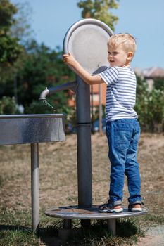 A happy little boy plays with a big water tap in a city park. Special water equipment for children's games on a hot summer day outdoors.
