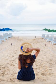 Beautiful girl in a hat sits with her back on a sandy beach and looks on sea.