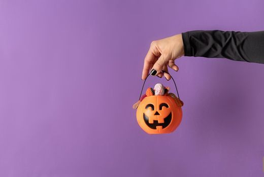 Happy Halloween concept. Scary woman Halloween hand with black nails holding pumpkin full of sweets on purple background with copy space