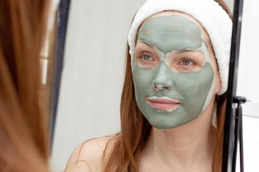 Smiling middle aged woman with band on head looking at her face with blue clay mask in mirror