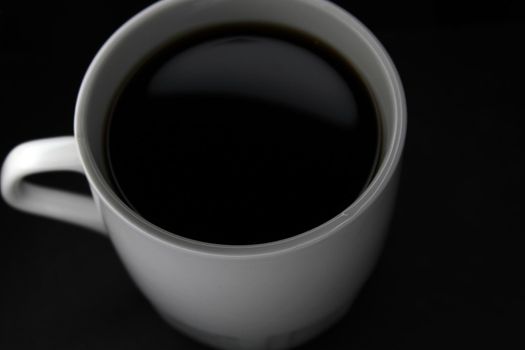 White mug with black coffee on a black background. View from above. Close-up..