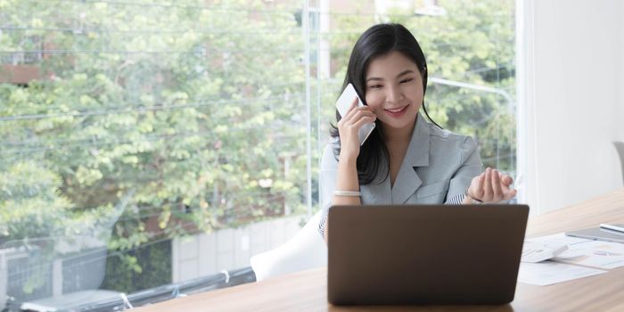 Attractive smiling young asian business woman relaxing at office, working on laptop computer, talking on mobile phone.