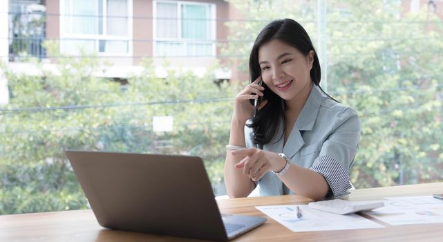 Attractive smiling young asian business woman relaxing at office, working on laptop computer, talking on mobile phone.