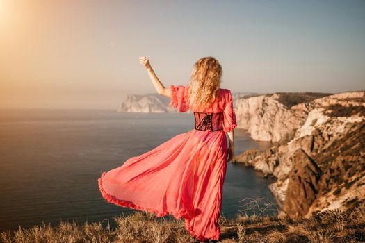 Side view a Young beautiful sensual woman in a red long dress posing on a volcanic rock high above the sea during sunset. Girl on the nature on blue sky background. Fashion photo