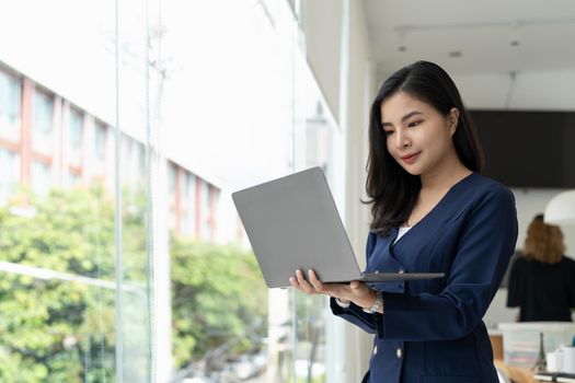 Asian businesswoman standing in modern office and using laptop computer.