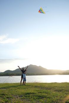 Children are flying kites while running on a meadow by the lake at sunset with their mother. Healthy summer activity for children. Funny time with family.