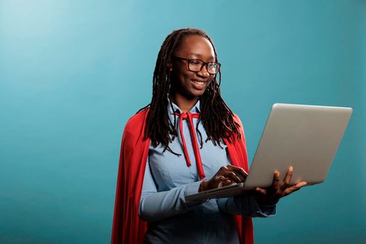 Brave and proud young adult superhero woman using modern laptop while standing on blue background. Confident and joyful justice defender wearing mighty hero cape while having handheld computer.