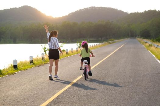 Asian mother teaching daughter to ride a bicycle in the park. A cute happy little girl learning to ride a bicycle with her mother. Active outdoor activities for kids.