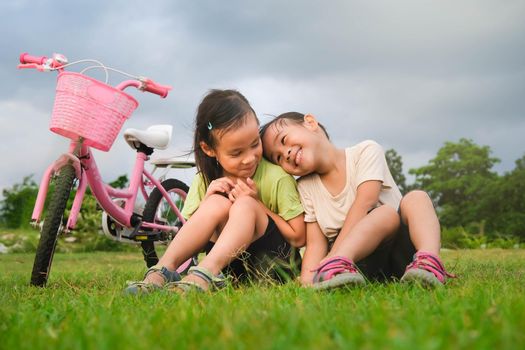 Happy cute little girl and her sister sitting on the lawn near the bikes in the park. Kids resting after biking. Healthy Summer Activities for Kids