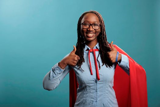 Positive smiling heartily justice defender wearing mighty hero red cape while giving approve sign. Young adult brave and confident looking superhero woman showing thumbs up gesture to camera