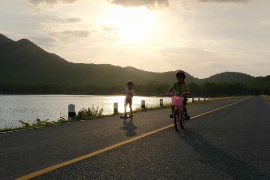 Cute little girl riding a bicycle and her sister riding a scooter on a lake road at sunset. Happy sisters doing outdoor activities together. Healthy Summer Activities for Kids.