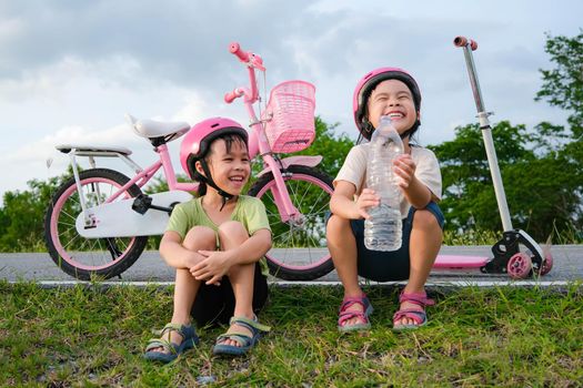 Happy cute little girl and her sister sit on the roadside lawn and drink water from a bottle near a bike in the park. Kids resting after biking. Healthy Summer Activities for Kids.