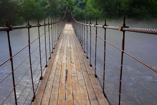 old wooden long suspension bridge over the Belaya mountain river, in nature, thick mystical fog over the water in the early morning