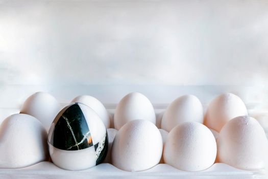 Eggs of different colors in cardboard boxes. A marble stone black-and-white egg stands in a cassette in the middle of white chicken eggs. The concept of contrast, individuality, exclusivity. concept of a damaged tooth