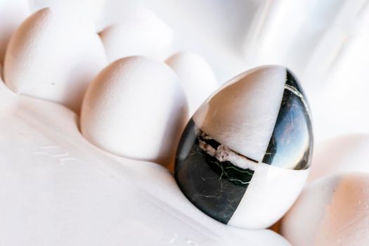 Eggs of different colors in cardboard boxes. A marble stone black-and-white egg stands in a cassette in the middle of white chicken eggs. The concept of contrast, individuality, exclusivity. concept of a damaged tooth