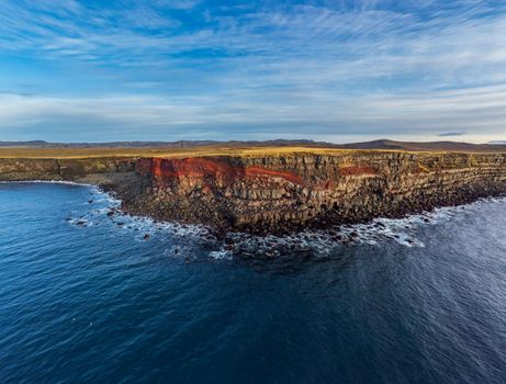 Wide angle oceans view of volcanic steep cliffs with red seams in Iceland