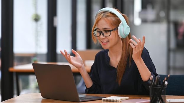 Young female employee wearing wireless headphone and watching online webinar on laptop computer.
