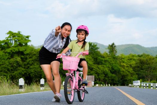 Asian mother teaching daughter to ride a bicycle in the park. A cute happy little girl learning to ride a bicycle with her mother. Active outdoor activities for kids.