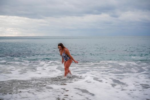 A plump woman in a bathing suit enters the water during the surf. Alone on the beach, Gray sky in the clouds, swimming in winter