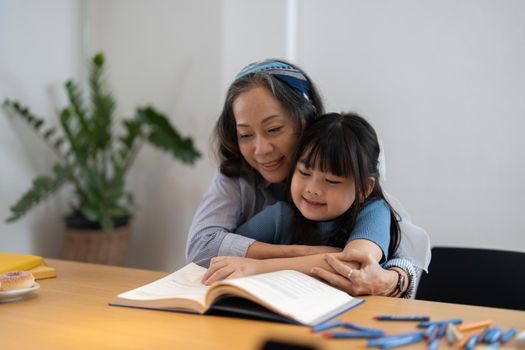 Close up smiling mature grandmother reading book to little granddaughter, telling interesting fairytale story, loving elderly woman hugging pretty preschool girl, relaxing on cozy couch at home.