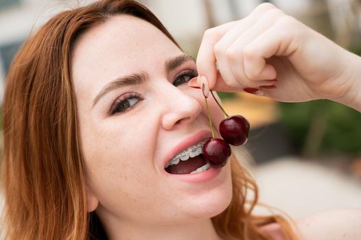 Beautiful young red-haired woman with braces on her teeth eats sweet cherries in the summer outdoors.