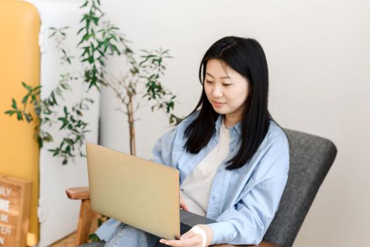 A young Asian woman works behind a laptop, sitting in the chair of the house. Korean woman enjoys online shopping from home