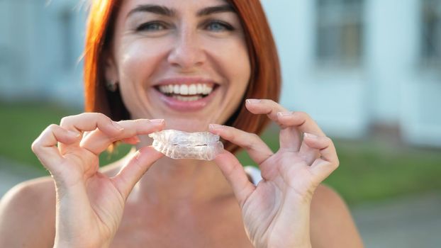 Red-haired Caucasian woman holding transparent mouthguards for bite correction outdoors. A girl with a beautiful snow-white smile uses silicone braces.